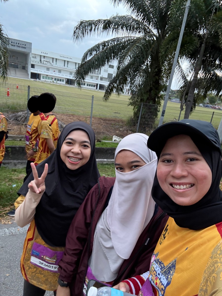 Three women are smiling whilst selfie. 2 male at the back, face covered.