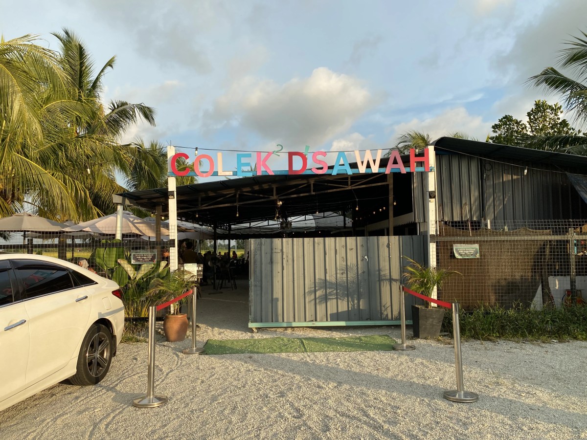 Entrance of Colek D'sawah restaurant with colorful signage, surrounded by palm trees and a white car parked nearby.
